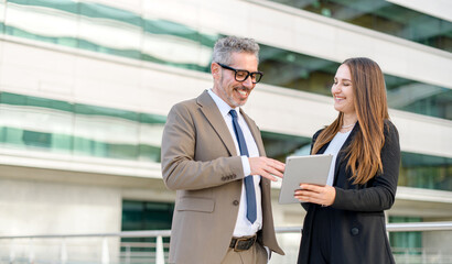 Experienced businessman with grey hair smiles as he looks at a tablet screen held by a young female...