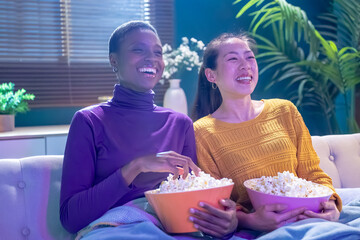 Two girl friends sitting on the couch watch a movie and eat popcorn. Funny friends relaxing together. High quality photo
