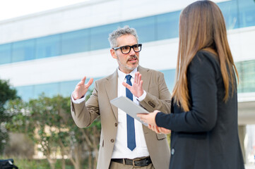 In a casual outdoor meeting, a grey-haired businessman animatedly explains a concept to a young female professional, their open body language indicative of constructive and dynamic business discussion