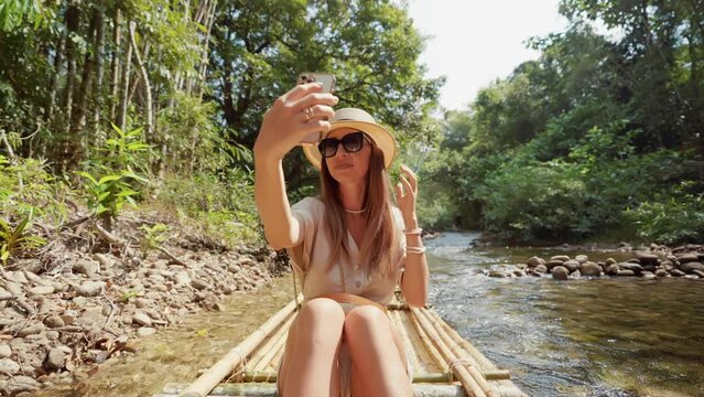 Female tourist floats on bamboo raft. Woman tourist enjoying the bamboo rafting on the river, films beautiful nature landscape on smartphone. Vacation, tropical tourism, travel, cheerful concept.
