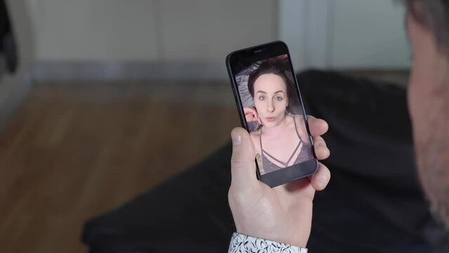 A middle aged man sat on a sofa talking to an attractive woman who is talking in her bedroom flirting on a smart phone over a video call, long distance work from home concept