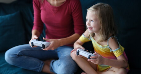 Little girl and mother sitting on couch and holding computer joysticks in their hands. Child addiction to computer games concept