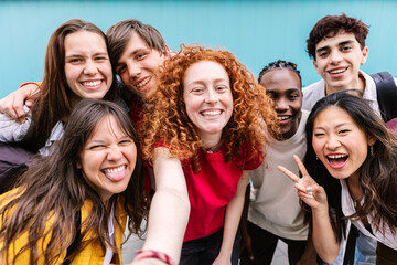 Diverse student friends group bonding together outdoors, taking selfie portrait standing over blue background. Youth community and teenage friendship concept. - obrazy, fototapety, plakaty