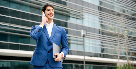 Confident smart Hispanic businessman in a blue suit happily converses on his smartphone while holding a laptop, showcasing flexibility and connectivity in the corporate world.