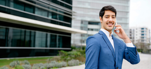 A dapper Hispanic businessman in a blue suit is engaged in a phone conversation, with a sleek office building highlighting themes of corporate communication and modern professionalism.