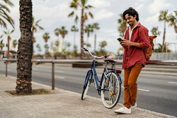 Portrait of a trendy man standing on city street with bike next to him and sending online messages.