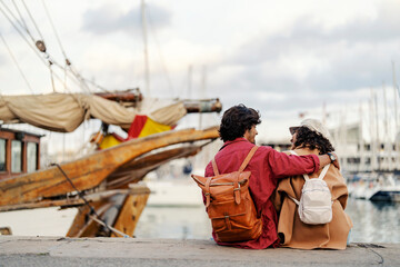 Rear view of a happy trendy young couple sitting at the pier near the ocean and hugging.