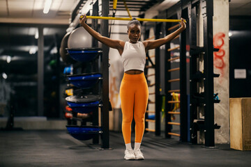 A slim black sportswoman is practicing her arms with resistance band at gym.
