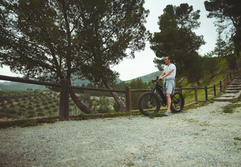 Obraz premium Athlete man takes a break while rides e-mountain bike on trail above olives valley in mountains