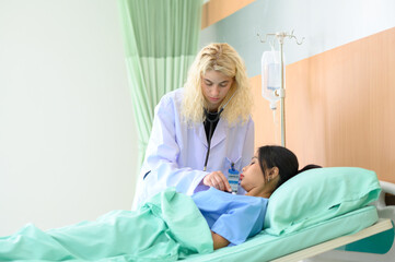 Young female doctor uses stethoscope to listen to heartbeat and lungs of recovering female patient resting in bed.
