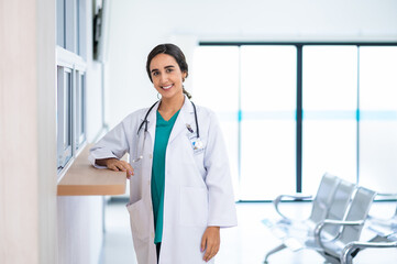 Profile photo of attractive smiling female doctor wearing white medical gown with stethoscope, standing in hospital corridor. Medicine and health care concept with copy space. looking at camera.