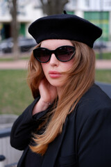 young blonde in a black beret and black glasses. woman in a beret and glasses, portrait photo of a blonde. Woman outdoors wearing sunglasses