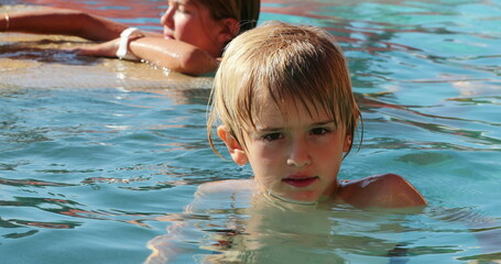 Portrait of blond small boy inside water at the swimming pool looking to camera