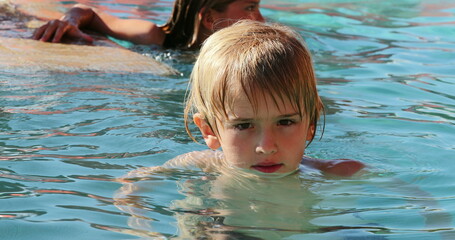 Portrait of blond small boy inside water at the swimming pool looking to camera