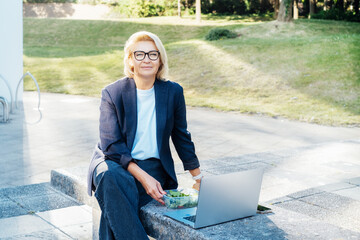 Business woman eating fast and healthy in front of laptop during job lunch break outdoors. Modern...
