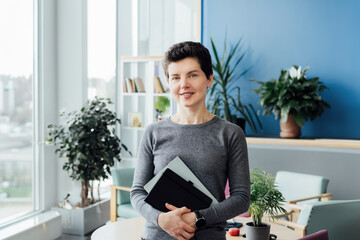 Portrait of smiling middle aged woman in casual outfit with digital tablet and notebook standing in...