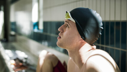 Profile of a pensive young swimmer looking at the distance while sitting at the poolside with...