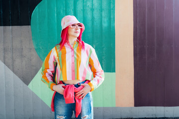 Young woman with pink hair and sunglasses in Bucket hat and multicolor strippled shirt posing on...