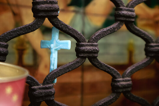 Inquadratura ravvicinata di un piccolo crocifisso azzurro appeso a una grata protettiva in ferro battuto di un'antica edicola votiva, simbolo di fede cristiana, speranza e devozione religiosa