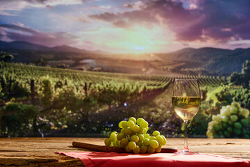 Glass of white wine and bunch of grapes on a wooden table. Blurred vineyard with rows of grapevine on background. Concept of winemaking, organic beverage, nature, traditions - obrazy, fototapety, plakaty