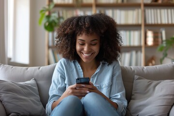 Happy girl checking social media holding smartphone at home. Relaxed smiling young woman sitting on couch using cellphone. Curly black hair teen female use mobile phone, playing game, shopping online - obrazy, fototapety, plakaty