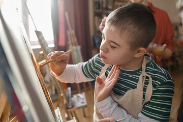 Close up portrait of young boy with Down syndrome painting on easel in sunlight enjoying art class