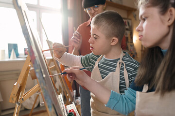 Side view portrait of children with disability painting together standing by easels in art class...