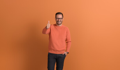 Young successful businessman in eyeglasses smiling and showing thumbs up sign on orange background