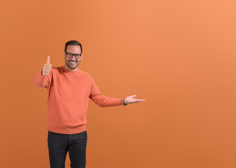 Happy businessman showing thumbs up sign and empty palm while sharing feedback on orange background