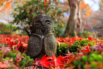 A Jizo statue with a smiling face is sitting on a pile of red leaves. - 779895007