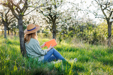 Woman is relaxing in blooming orchard and reading book to improve her mindfulness and mental health. Digital detox outdoors at springtime