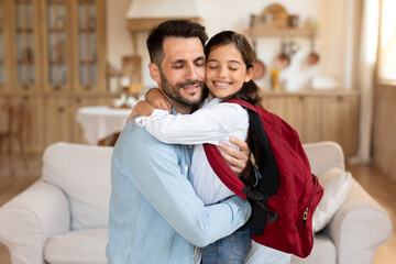 Father embracing schooler daughter with backpack at home