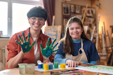 Front view portrait of young girl with disability smiling at camera in art studio with cheerful teacher showing hands covered in paint - obrazy, fototapety, plakaty