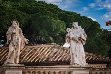 Obraz premium Weathered statues of a bearded figure and a figure holding a child on a Mediterranean tiled roof with lush green trees in the background. Likely in the Vatican.