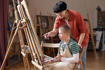 Side view portrait of smiling female teacher assisting boy with down syndrome painting on easel in art class