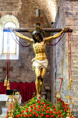 A statue of Jesus Christ is holding a cross in his hands. The cross is decorated with red beads and...
