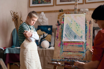 Side view portrait of anxious young boy with Down syndrome standing by easel in art class and...