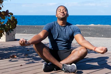 Poster Young happy man practicing yoga by seaside on sunny day with friends, enjoying warmth of sunshine with smiling face while sitting in lotus position and meditating. Sport during vacation © neirfy