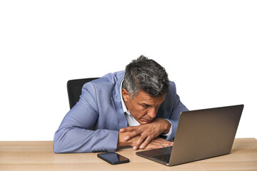 Tired middle-aged Latino man leaning on table while working on laptop