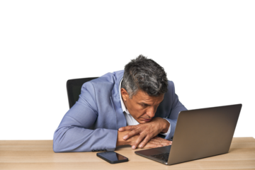 Tired middle-aged Latino man leaning on table while working on laptop