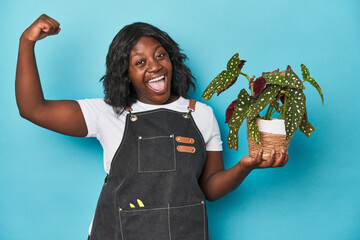 Curvy gardener holding plant in studio raising fist after a victory, winner concept.