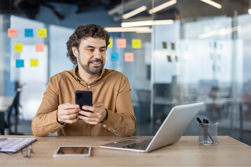 Professional hispanic man with a mature smile using a smartphone in a modern office environment,...