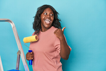 Curvy woman with paint roller and ladder points with thumb finger away, laughing and carefree.