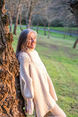 A smiling middle-aged woman leaning against a tree trunk in the park.