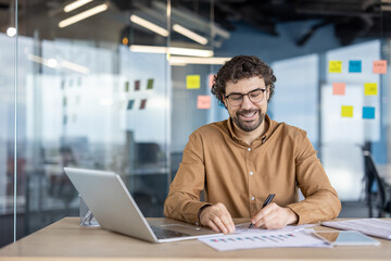 Happy mature Hispanic man enjoying his work with laptop and documents in a modern office setting. Positive work environment and job satisfaction concept.