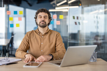 A male employee uses breathing techniques to find calm and focus at his office workspace, eyes...