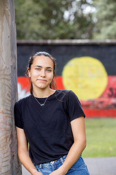 Aboriginal woman leaning against a gumtree in front of an Aboriginal flag mural in a park