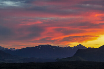 Scenic view of snow capped mountain peak Dobratsch at sunset seen from Taborhoehe in Carinthia, Austria, Europe. Sky has vibrant orange and pink colors with clouds swirling around summit. Serenity