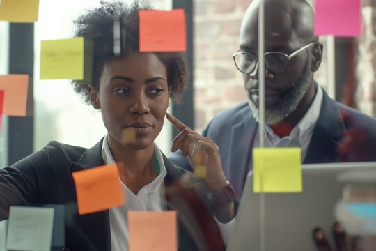 Young mixer race businesswoman manager explaining strategy ideas on sticky notes on glass wall to male African American colleague looking at strategy scrum presentation. Business project planning