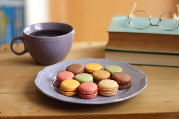 Purple plate filled with pastel macarons, cup of tea or coffee, vintage books and reading glasses on the table. Colorful bookcase in the background. Selective focus. 
